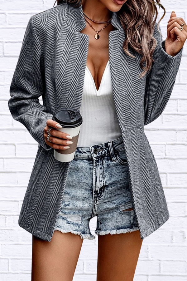 Person wearing a gray blazer, white top, and denim shorts holding a coffee cup against a white brick wall.